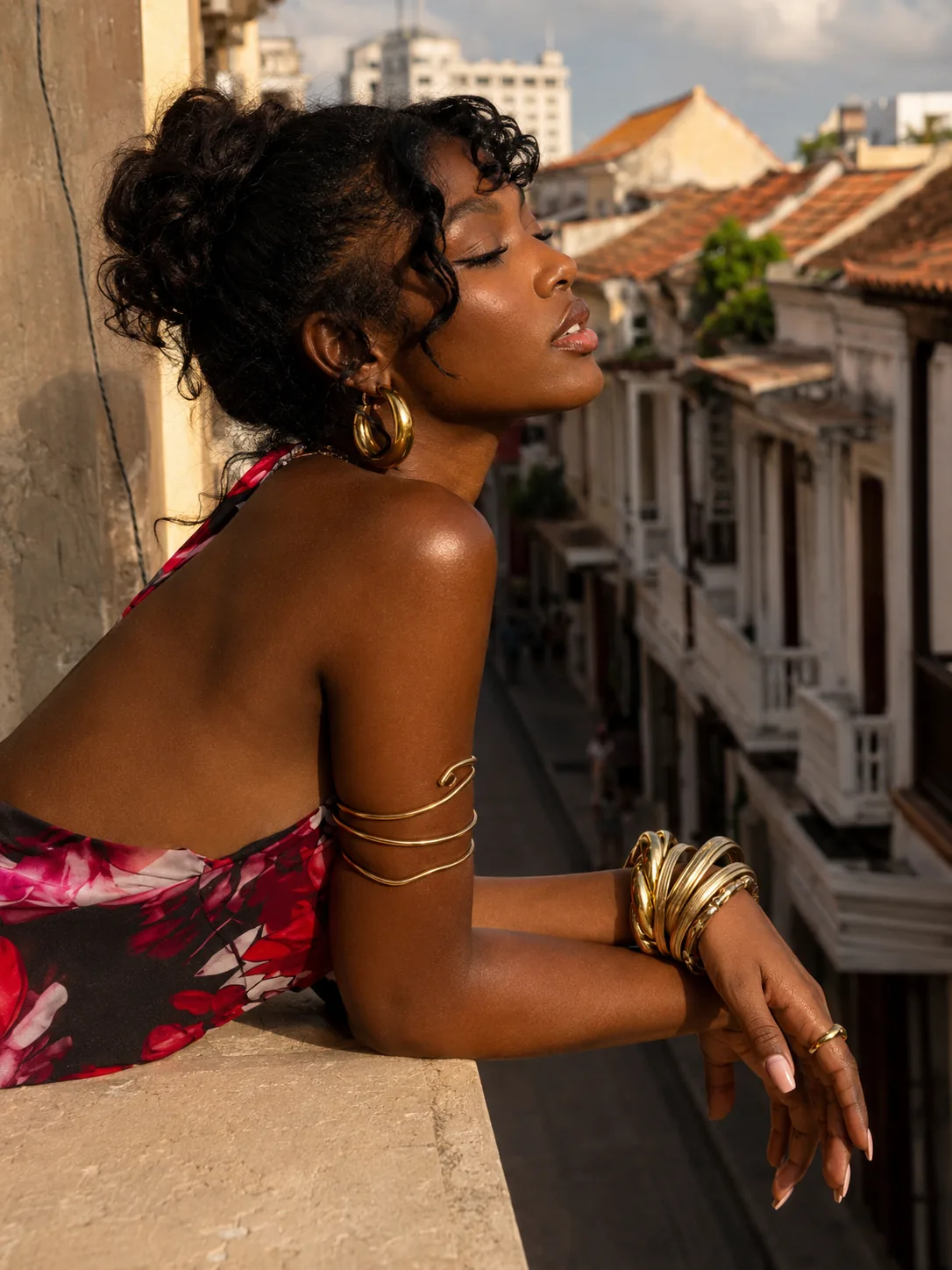 Editorial beauty portrait of a woman on a sunlit balcony with glossy lips and gold jewelry.